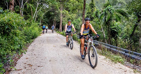 A group of people riding e-bikes on a gravel path through lush greenery, enjoying an outdoor cycling adventure in a natural environment.